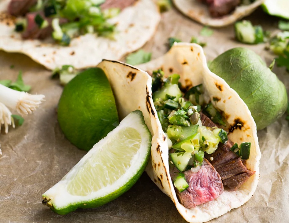 meat taco with avacado topping surrounded by ingredients including lemons